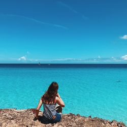 Rear view of woman sitting on shore against blue sea