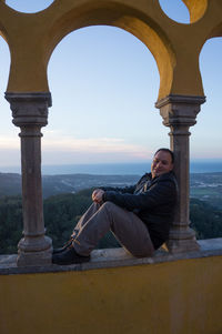 Side view of man sitting on retaining wall against sky