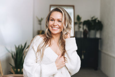 Blonde smiling woman 35 year with long hair in cozy knitted cardigan at the bright interior