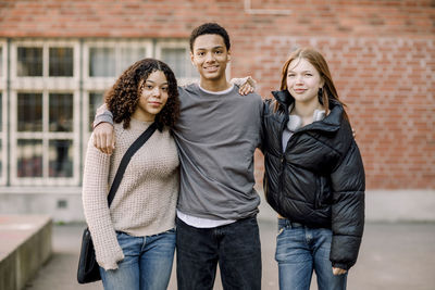 Portrait of smiling male teenage boy standing with arms around female friends in high school campus