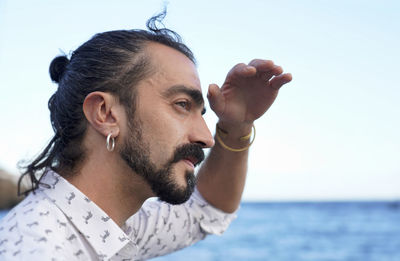 Profile view of mid adult man shielding eyes while standing at beach against sky