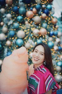 Portrait of smiling young woman standing by christmas tree holding cotton candy outdoors