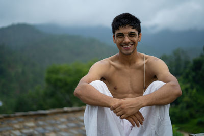 Young shredded man sitting wearing a dhoti and glasses with blurred mountains in the background.