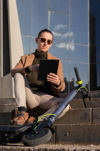 Vertical smiling young man using a tablet sitting in the financial district with electric scooter