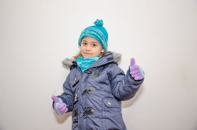 Portrait of cute girl wearing warm clothing showing thumbs up sign against wall