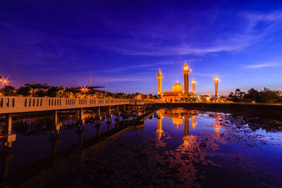 Illuminated building by river against sky at sunset