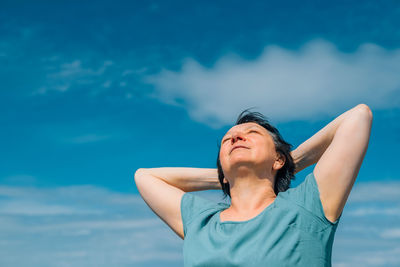 Close-up portrait of an adult smiling woman with her hands behind her head against a blue sky 