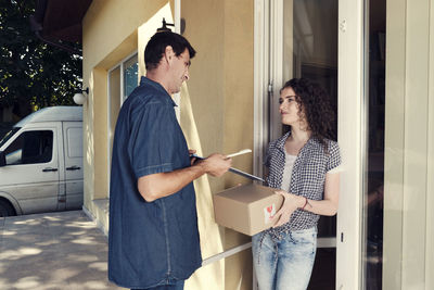 Delivery man giving pen and clipboard to woman against house