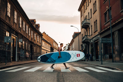 Man walking on crosswalk holding paddleboard in hand against sky