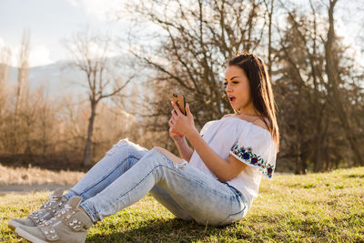 Shocked woman using mobile phone while sitting on field