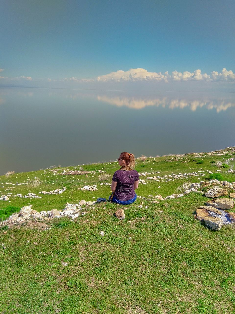 sky, beauty in nature, real people, leisure activity, one person, rear view, lifestyles, scenics - nature, cloud - sky, nature, tranquil scene, day, tranquility, land, casual clothing, non-urban scene, grass, sitting, plant, water, outdoors