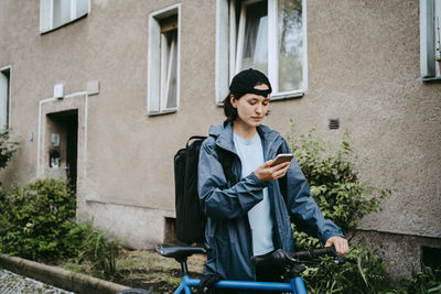 Female biker using smart phone while standing with bicycle near building