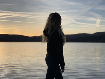 Woman standing by lake against sky during sunset