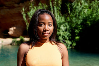 African american female traveler in casual clothes looking at camera while standing on shore of clean lake against cliff with waterfall on summer weekend day