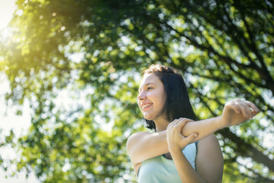 Low angle view of smiling teenage girl looking away while exercising against trees at park