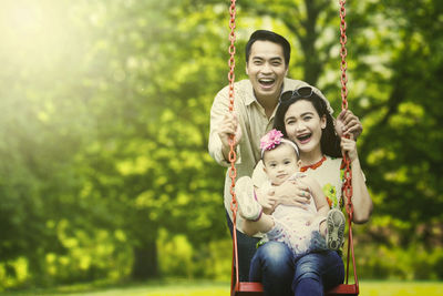 Portrait of happy parents with daughter sitting on swings against trees in park