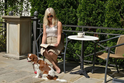 Blonde woman sitting in elegant garden with her pet, reading a book and drinking coffee