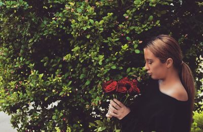 Side view of young woman holding roses while standing by plants at park