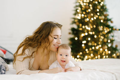 Happy mother lies on the bed next to her baby in the background of christmas lights