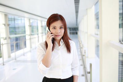 Portrait of young woman holding disposable cup while standing by window in office
