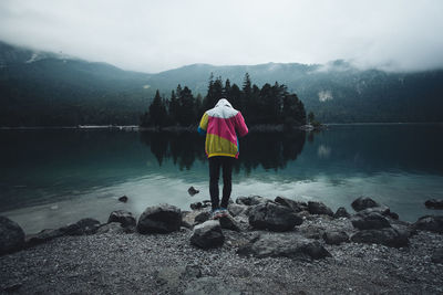 Rear view of man standing on rock at lakeshore against mountains