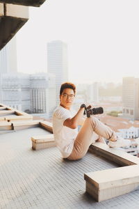 Side view of young man holding digital camera while sitting on floor in city