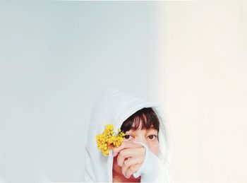 Close-up portrait of woman in hooded shirt holding flower against white wall