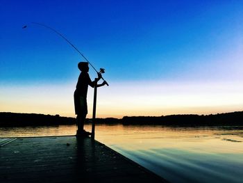 Boy with fishing rod standing by river against blue sky