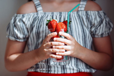 The hands of a girl in a striped blouse and a red skirt hold a glass with juicy strawberries.