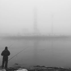 Full length rear view of man fishing at lakeshore during foggy weather