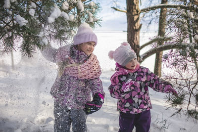 Happy girl friends enjoy winter season and pour snow from trees in forest on sunny winter day