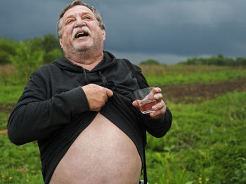 Happy senior man with whiskey glass showing abdomen on field