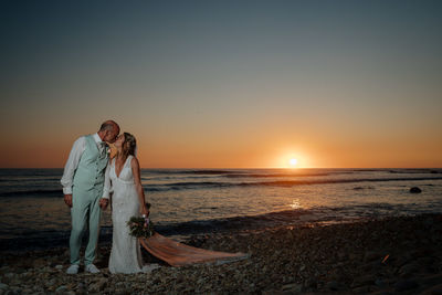 Bride and groom at beach against sky during sunset