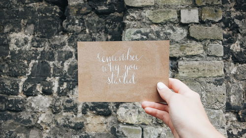 Cropped image of hand holding paper with text against wall