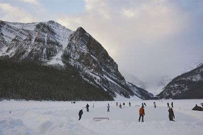 People skiing on frozen lake louise in front of mountains against cloudy sky