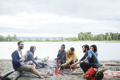 Happy friends roasting sausages on campfire at riverbank against sky