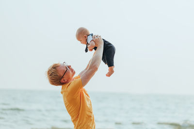 Side view of father holding baby against sea and sky