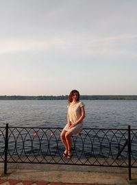 Woman sitting on metallic railing at promenade by lake against sky