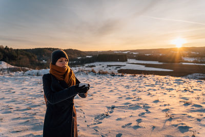 Young woman in coat and hat at sunset stands on snow-covered field, holds handful of snow in hands
