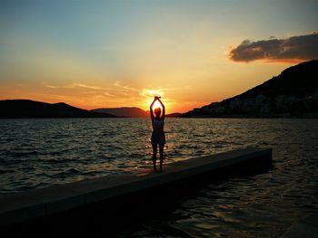 Silhouette woman with arms raised standing amidst river against sky during sunset