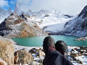 Low section of person sitting against lake and snowcapped mountains during winter