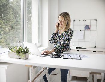 Smiling businesswoman talking on phone while using laptop in office