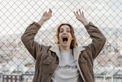 Young beautiful woman screaming looking at camera in front of metal fence with city view behind