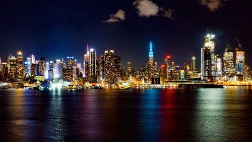 Illuminated modern buildings by sea against sky at night