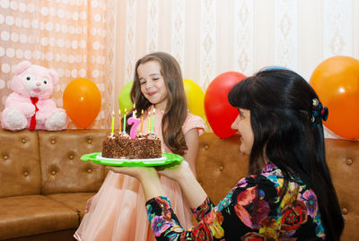 Mature woman holding birthday cake with lit candles by happy daughter at home