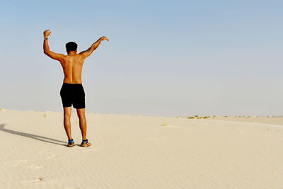 Rear view of shirtless man with arms raised standing at desert against clear sky