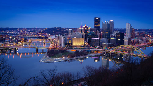 Illuminated bridge over river by buildings against sky at dusk