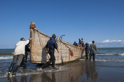 People standing by boat on shore at beach against blue sky