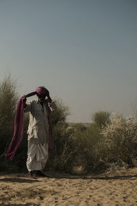 Man tying turban while standing on sand at desert against clear sky during sunny day