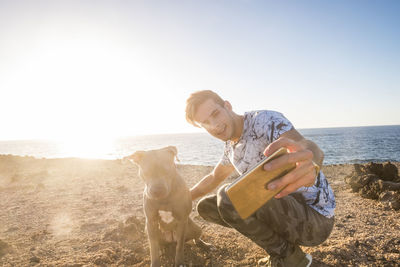 Smiling young man taking selfie with dog at beach during sunset